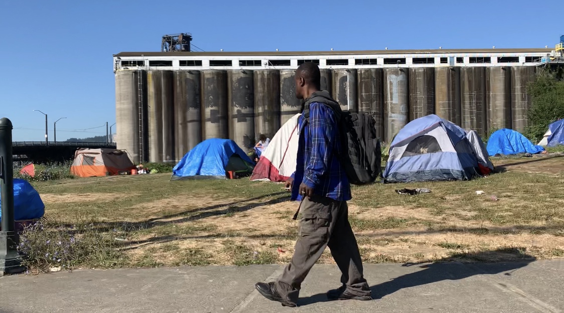 A man walks past a homeless camp in Portland along the Willamette River directly across from the Moda Center, 2020.