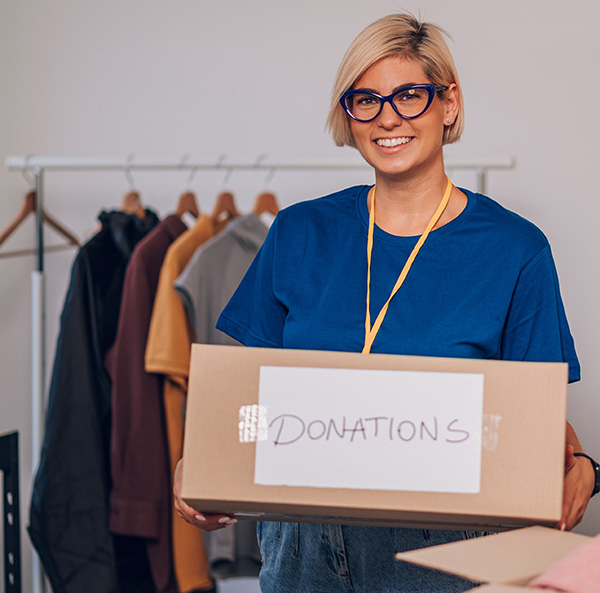 Portrait of a young woman working in charitable foundation and posing while holding box in her hands. Female volunteer in blue uniform looking into the camera while helping in a donation center.